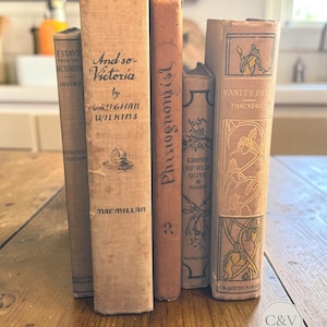 Peut inclure: Une collection de cinq livres reliés anciens disposés verticalement. Les dos présentent des titres tels que Essays from the Sketch Book, And so-Victoria de Vaughan Wilkins, Physiognomist, Crown of Wild Olive de Ruskin et Vanity Fair de Thackeray.