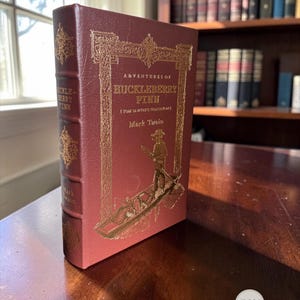 Pode incluir: Um livro de capa dura cor de vinho intitulado Adventures of Huckleberry Finn de Mark Twain. A capa apresenta estampagem em folha de ouro com uma borda ornamentada e um gráfico de uma pessoa num barco. A lombada também exibe letras douradas.