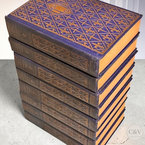 May include: A stack of seven hardcover books with purple covers featuring a gold diamond lattice pattern and fleur-de-lis motifs. The spines are dark purple with gold geometric detailing and text. The page edges are a solid orange color.