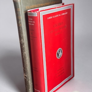 May include: Two hardcover books standing upright. The front book is bright red with white text reading Loeb Classical Library, Cicero On The Orator Books 1-2. The back book is dark olive green with gold text reading Gildersleeve-Lodge Latin Series.