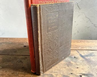 以下が含まれることがあります： 直立した2冊のヴィンテージハードカバー本。後ろの本は赤色で、背表紙にThomas Carlyleの文字があります。前の本は茶色で、質感のある表紙にCarlyle Essay on Burnsの文字が前面パネルに印刷されています。
