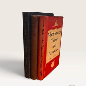 Puede incluir: Tres libros de tapa dura antiguos titulados Mathematical Tables and Formulae de F.J. Camm. Los libros están dispuestos en fila, con un volumen negro, uno marrón y uno rojo con un panel de cubierta color canela y una ilustración de triángulo.