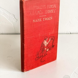 以下が含まれることがあります： マーク・トウェイン著『Extracts From Adam's Diary』の赤いハードカバー本。表紙には金色のエンボス加工されたタイトルと、石板に文字を書く人物と足元に座る2匹の小さな動物を描いた黒い線画が施されています。