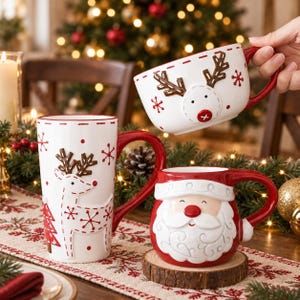 Könnte beinhalten: Drei Keramiktassen mit weihnachtlichen Motiven. Eine hohe weiße Tasse zeigt ein Rentier und rote Bäume. Eine zweite weiße Tasse zeigt ein Rentiergesicht mit Geweih. Eine dritte rote Tasse stellt ein Weihnachtsmann-Gesicht mit weißem Bart dar.