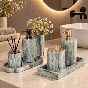 Può includere: Un set di accessori da bagno in marmo verde, composto da due dispenser per sapone con erogatori in metallo, un contenitore con coperchio, un vaso cilindrico e un diffusore a bastoncini. Gli oggetti sono disposti su due vassoi in marmo coordinati.