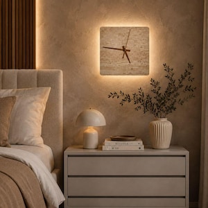Puede incluir: Un reloj de pared cuadrado con un acabado de piedra beige texturizada y manecillas finas de color marrón oscuro. El reloj está montado en la pared y cuenta con una luz de fondo cálida y suave que ilumina los bordes del marco cuadrado.