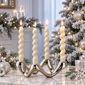 May include: A silver-toned metal candle holder with a wavy, fluid design holding four lit, off-white spiral taper candles. The candle holder features a smooth, reflective surface and is positioned upright to support the four vertical candles.