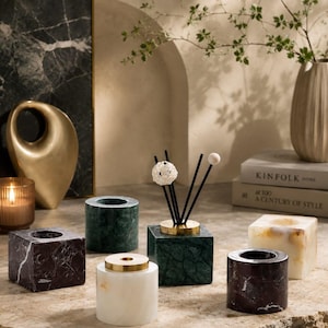 Puede incluir: Una colección de portavelas de piedra y una base para difusor de varillas en varias formas y colores, incluyendo mármol verde oscuro, negro y blanco. El conjunto presenta recipientes cilíndricos y cúbicos con detalles en tono dorado metálico.