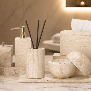 May include: A bathroom accessory set crafted from beige travertine stone, including a toothbrush holder, two soap dispensers with gold pumps, a tissue box cover, a lidded jar, and a reed diffuser holder with three black reeds.