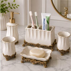 Op de afbeelding: Een badkameraccessoireset bestaande uit een crèmekleurige keramische zeepdispenser, een tandenborstelhouder met twee tandenborstels en een tube Sensodyne-tandpasta, een zeepbakje met een stuk zeep en een beker, allemaal met sierlijke gouden voetjes.