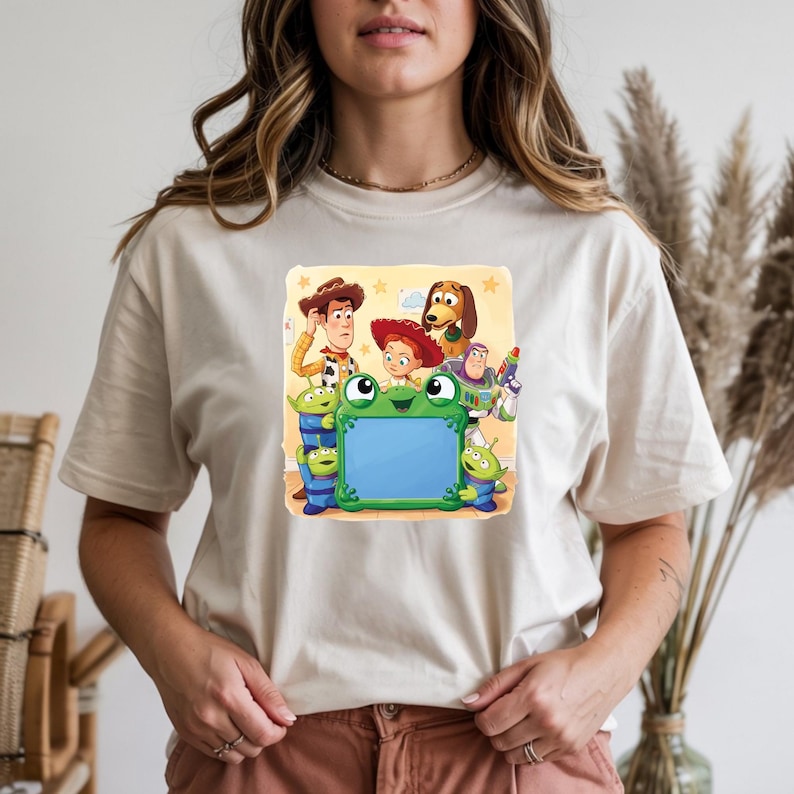 May include: A beige short-sleeve t-shirt featuring a graphic print of Toy Story characters including Woody, Jessie, Buzz Lightyear, Slinky Dog, and three green aliens surrounding a large, central, bright green frog-shaped picture frame.