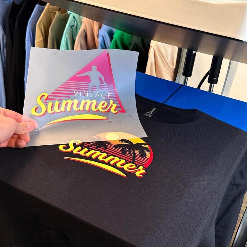 May include: A black t-shirt being printed with a heat transfer design. The transfer sheet shows a pink triangle with a silhouette of a person and the text "VINTAGE Summer". The shirt features a printed graphic of a sunset, palm trees, and "Summer".