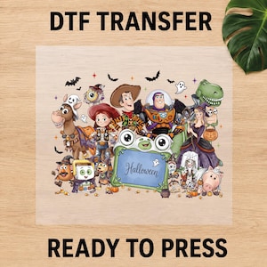 May include: A DTF transfer sheet featuring a group of cartoon characters dressed in Halloween costumes, including a cowboy, space ranger, cowgirl, and dinosaur. A central green frog-shaped frame displays the text Halloween in cursive script.
