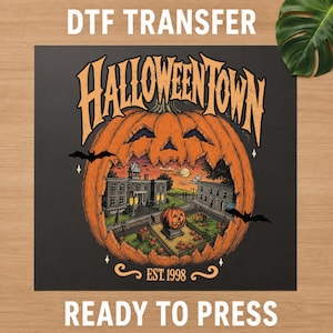 May include: A DTF transfer sheet featuring a large orange jack-o'-lantern with the text HALLOWEENTOWN and EST. 1998. Inside the pumpkin is a detailed illustration of a town square with buildings, a smaller pumpkin statue, and flying bats.