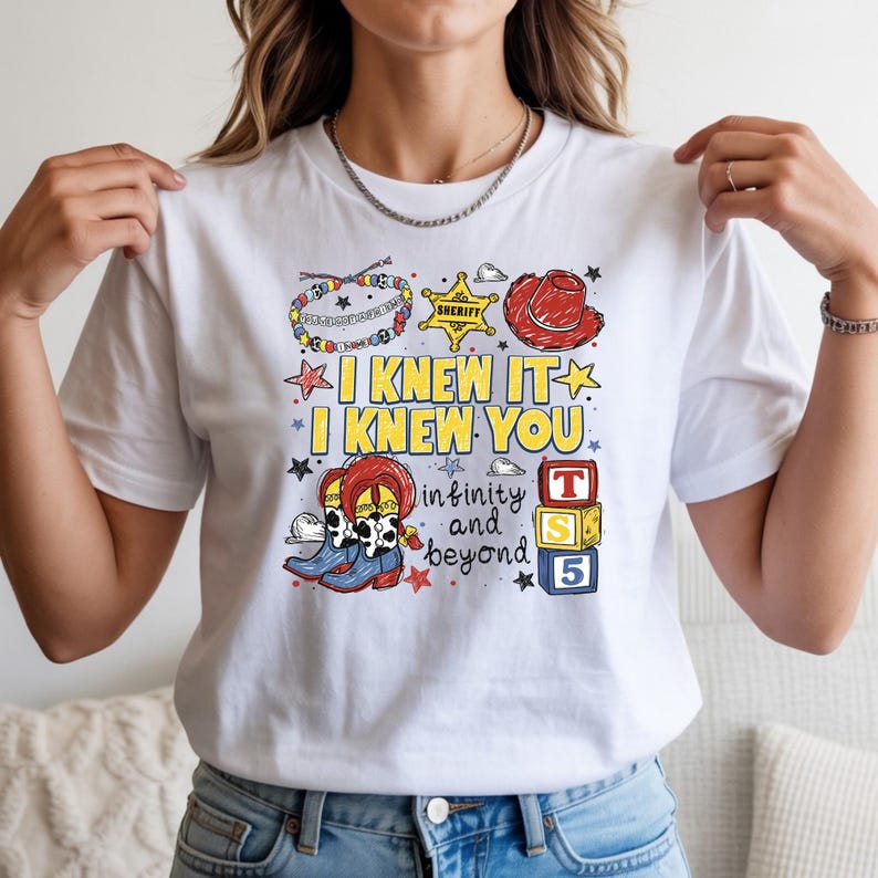 May include: A white t-shirt featuring a colorful graphic print with the text I KNEW IT I KNEW YOU and infinity and beyond. The design includes a beaded bracelet, a yellow sheriff star, a red cowboy hat, cowboy boots, and stacked toy blocks.
