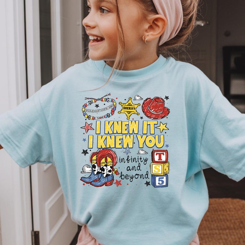 May include: A light blue t-shirt featuring a graphic print with the text I KNEW IT I KNEW YOU and infinity and beyond. The design includes a beaded bracelet, a yellow sheriff star, a red cowboy hat, cow-print boots, and three stacked blocks.