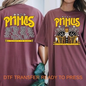 May include: A mauve t-shirt featuring a yellow Primus logo and A Tribute to Kings text. The back displays a list of tour cities, while the front shows a graphic of a crowned figure with swords and wings in black, white, and yellow.