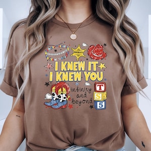 May include: A brown t-shirt featuring a graphic print with the text "I KNEW IT I KNEW YOU" and "infinity and beyond". The design includes a sheriff badge, a red cowboy hat, a beaded bracelet, cowboy boots, and three stacked toy blocks labeled T, S, 5.