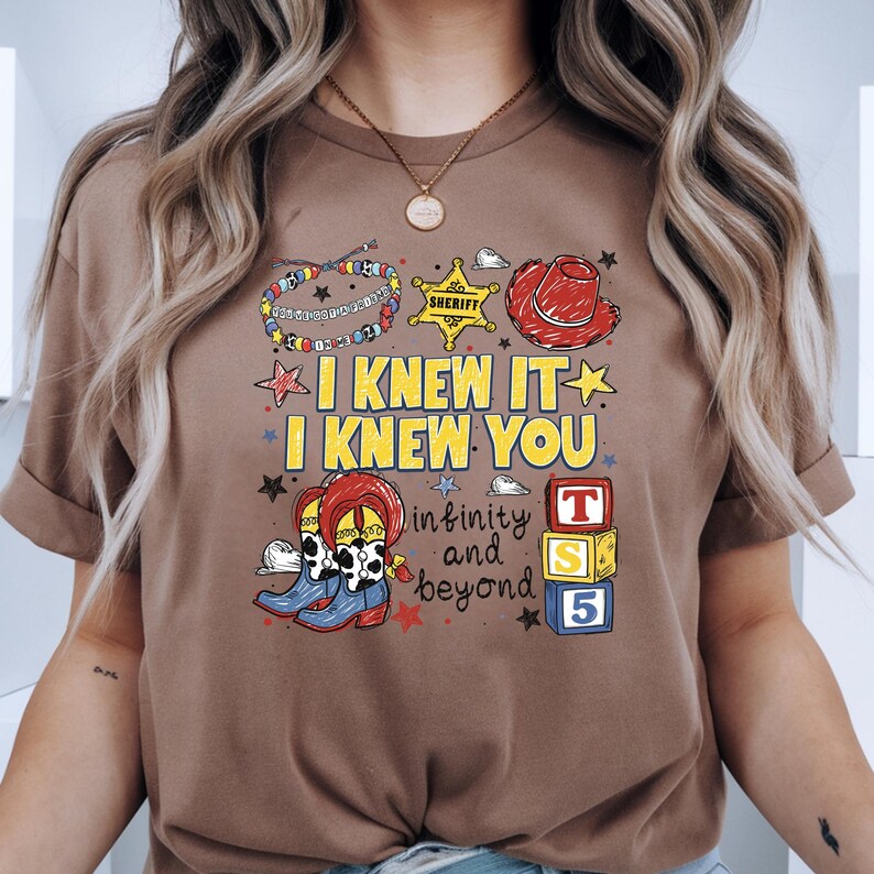 May include: A brown t-shirt featuring a graphic print with the text "I KNEW IT I KNEW YOU" and "infinity and beyond". The design includes a sheriff badge, a red cowboy hat, a beaded bracelet, cowboy boots, and three stacked toy blocks labeled T, S, 5.