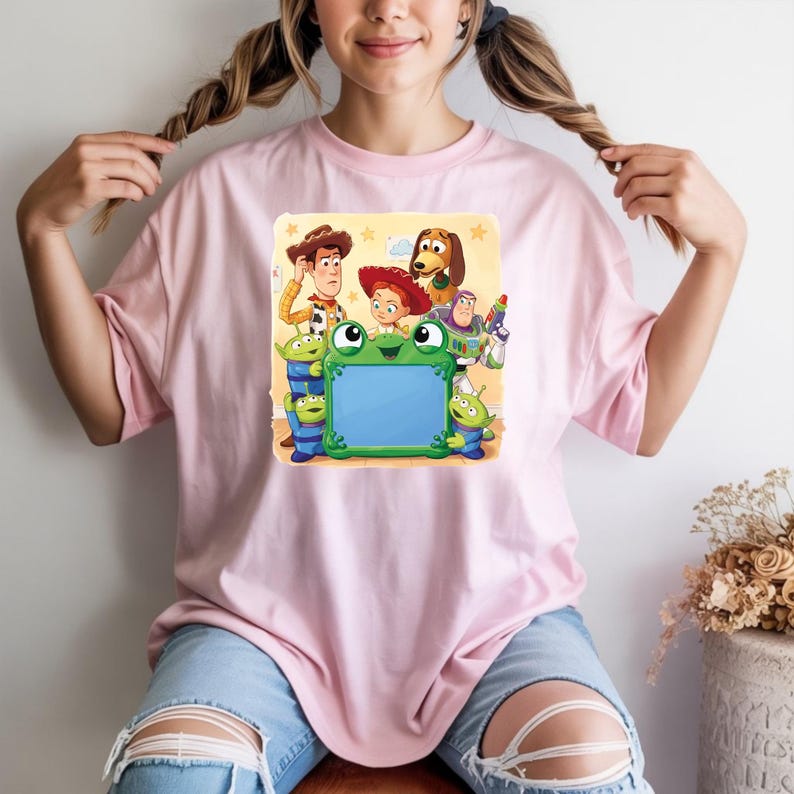 May include: A light pink short-sleeved t-shirt featuring a graphic print of Toy Story characters including Woody, Jessie, Buzz Lightyear, Slinky Dog, and three green aliens surrounding a large, empty, rectangular green frog-shaped frame.