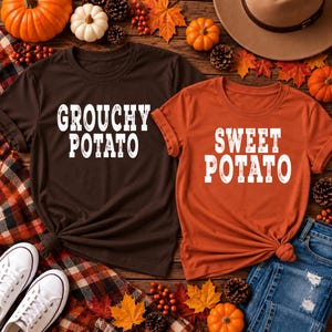 May include: A dark brown t-shirt with the white text GROUCHY POTATO and an orange t-shirt with the white text SWEET POTATO. Both shirts feature short sleeves and a crew neckline, presented as a pair of matching garments for casual wear.