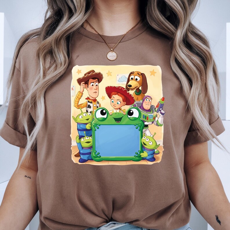 May include: A brown t-shirt featuring a colourful graphic print of Toy Story characters including Woody, Jessie, Buzz Lightyear, Slinky Dog, and three green aliens surrounding a large, central, green frog-shaped frame with a blank blue centre.