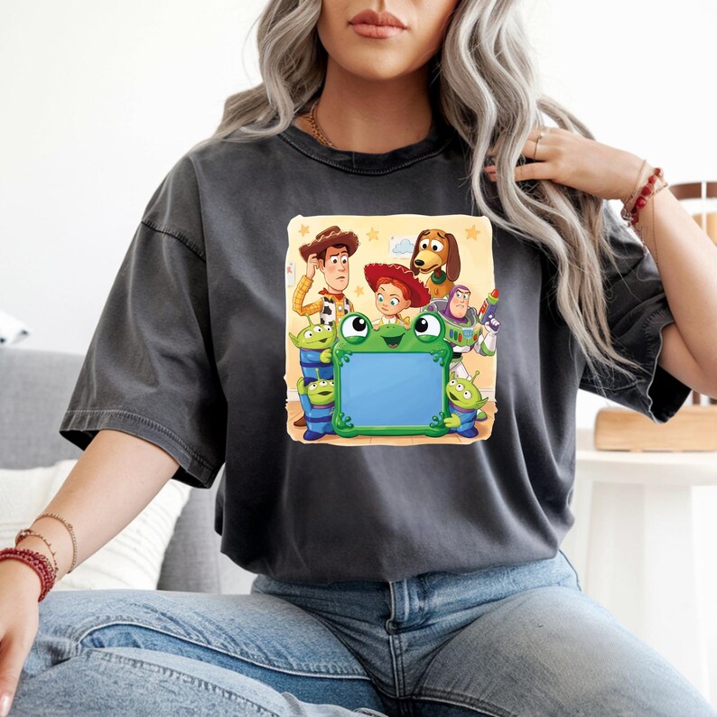 May include: A charcoal grey short-sleeve t-shirt featuring a graphic print of various animated characters surrounding a central green frog-shaped frame. The characters include a cowboy, a cowgirl, a space ranger, a dog, and three green aliens.