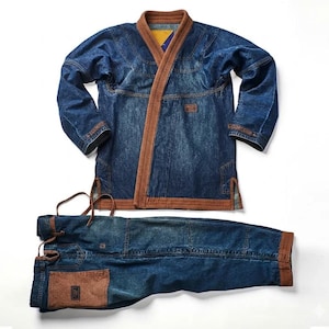Op de afbeelding: Een blauwe denim vechtsport gi-set bestaande uit een jas met lange mouwen en een bijpassende broek. De jas heeft een bruine geribbelde kraag en afwerking, terwijl de broek een bruine zak en een taille met trekkoord heeft.