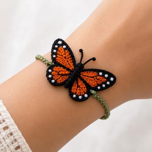 May include: A bracelet featuring a crocheted monarch butterfly with vibrant orange wings, black borders, and small white dots. The butterfly is attached to a thin, sage green braided cord that wraps around the wrist, showcasing detailed needlework.