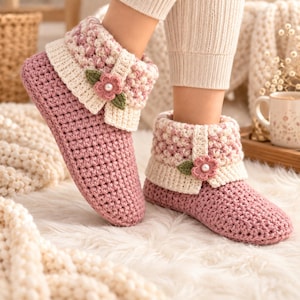Op de afbeelding: Een paar mauvekleurige gehaakte pantoffels met een textuurrijke, crèmekleurige boord. Elke pantoffel is versierd met een klein, gehaakt roze bloemetje met groene blaadjes en een centrale witte parel, wat een decoratief accent aan het schoeisel geeft.