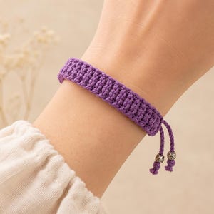May include: A purple woven cord bracelet featuring a textured, ribbed pattern. The bracelet includes two adjustable dangling ends, each finished with a small, metallic silver-toned bead with intricate engraved detailing at the very bottom.