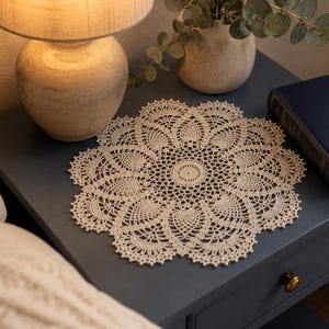 May include: A round, cream-colored crochet doily featuring an intricate, repeating floral-inspired pattern with a central circular motif and scalloped edges, crafted with fine thread to create a delicate, openwork design with a textured appearance.