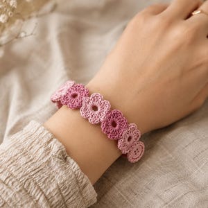 May include: A crochet bracelet featuring a series of interconnected flower motifs in varying shades of pink, ranging from light pastel to deep magenta. The bracelet is secured with a small, matching pink circular button closure at the end.
