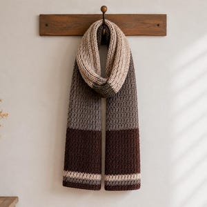 May include: A long, thick, crocheted scarf featuring a color-blocked design in shades of cream, light gray, medium gray, and dark brown. The scarf has a textured, ribbed stitch pattern and is draped over a hook on a wooden wall-mounted rack.