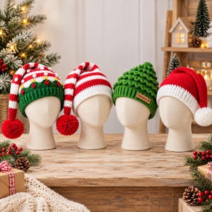 May include: Four crochet hats displayed on mannequin heads. The first is white and red with green trim and pom-poms. The second is red and white striped with a red pom-pom. The third is green with a textured surface and a small brown tag. The fourth is red with a white brim and pom-pom.
