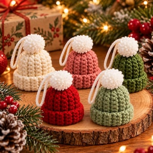Op de afbeelding: Vijf kleine, gehaakte mutsjes als ornament in crème, oudroze, donkergroen, felrood en saliegroen. Elke muts heeft een dikke geribbelde rand, een getextureerde kroon, een witte pompon bovenop en een wit garen lusje om op te hangen.