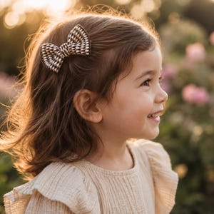 May include: A hair clip featuring a small, structured bow with a woven houndstooth pattern in shades of dark brown, black, and white. The bow is attached to a metal clip mechanism, designed to secure hair in place with a classic, textured finish.