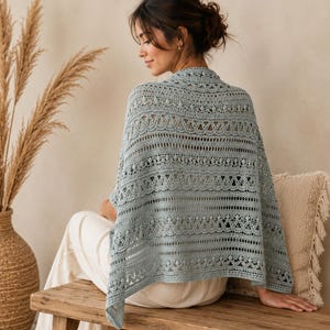 May include: A light blue crochet shawl featuring an intricate pattern of alternating openwork lace, solid bands, and small bobble stitches. The garment is draped over the shoulders, showcasing a detailed texture and a soft, muted colour palette.