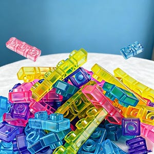 May include: A collection of translucent plastic building blocks in vibrant shades of pink, yellow, blue, green, and purple. The rectangular bricks feature circular studs on top and are scattered in a pile, with two individual pieces floating above.