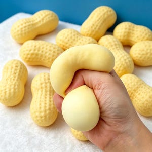 May include: A collection of pale yellow, textured peanut-shaped stress relief toys. One of the toys is being squeezed, showing its soft, pliable material and ability to deform under pressure, while the others remain in their original, firm shape.