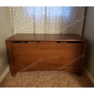 May include: A rectangular wooden storage chest with a medium brown finish and a smooth, flat top surface. The front panel features a solid construction with two semi-circular cutouts along the top edge to facilitate opening the hinged lid.