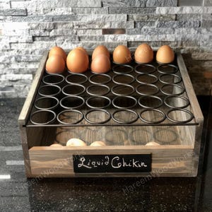 May include: A wooden egg holder with a metal wire grid for storage. The front features a black rectangular chalkboard label with the white handwritten text Liquid Chikin. Several brown eggs are placed in the top wire slots and bottom tray.