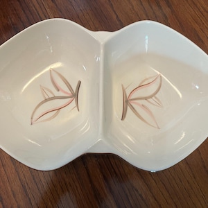 May include: A white ceramic divided serving dish with a symmetrical, leaf-like shape. Each of the two sections features a delicate, hand-painted botanical motif consisting of thin, stylized leaves in shades of soft pink and muted brown.
