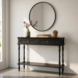 May include: A black console table with two drawers, brass knobs, and a lower shelf, topped with a light green vase containing dried stems, two stacked books titled The Architecture of Place and Naturalist, and a small potted succulent plant.