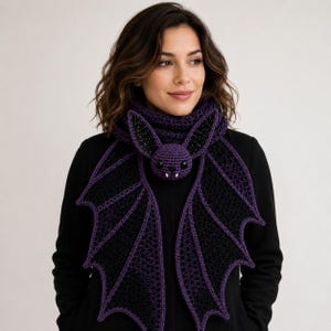 May include: A crochet scarf featuring a central purple bat head with black eyes and white fangs, connected to two large, intricate black and purple bat wings that drape downwards. The scarf is crafted with a textured, open-work stitch pattern.