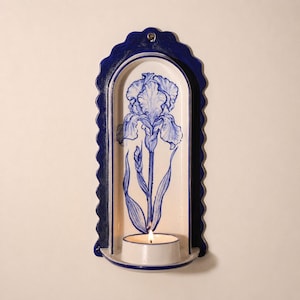 Puede incluir: Un portavelas de pared de cerámica con un marco festoneado azul oscuro y un panel trasero arqueado de color crema. El centro muestra una ilustración pintada a mano de una flor de iris azul. Una pequeña vela de té blanca está encendida.