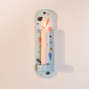 Peut inclure: Un bougeoir mural bleu clair de forme rectangulaire arrondie, orné d'un motif de coquillages colorés et de petites étoiles. Une bougie effilée rose pâle est allumée et repose dans le support intégré situé à la base de l'objet.