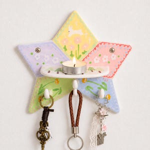 May include: A star-shaped ceramic wall hook rack with a small shelf holding a tea light candle. The surface features pastel-coloured sections with painted flowers, a white rabbit, and green trees. Three white hooks hang from the bottom holding keychains.