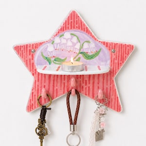May include: A star-shaped ceramic wall organiser in coral pink with vertical red stripes. It features a small shelf holding a lit tea light candle, a painted lily of the valley floral design on a lavender background, and three hooks holding keychains.