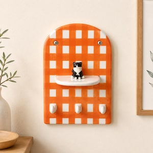 May include: An arched wall-mounted organiser featuring an orange and white gingham pattern. It includes a small white shelf with a black and white cat figurine sitting on it, and three white hooks at the bottom for hanging items, secured by two screws.